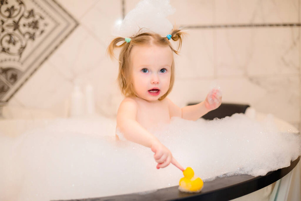 Menina bonito bebê tomar banho no banho com espuma em casa, brinca com pato de borracha  - Foto, Imagem
