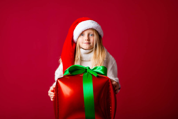 Happy little blonde girl in Santa's hat holding a Christmas gift. Copy space photo on isolated red background, New Year's sales concept - Foto, Imagem