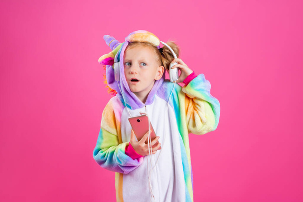 Feliz niña rubia en unicornio unitard escucha música desde su teléfono inteligente. Aislado sobre fondo rosa con espacio para texto - Foto, Imagen