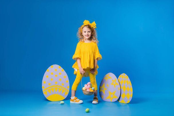 Sarı elbiseli küçük sarışın kız mavi arka planda büyük dekoratif paskalya yumurtalarıyla ayakta duruyor. - Fotoğraf, Görsel