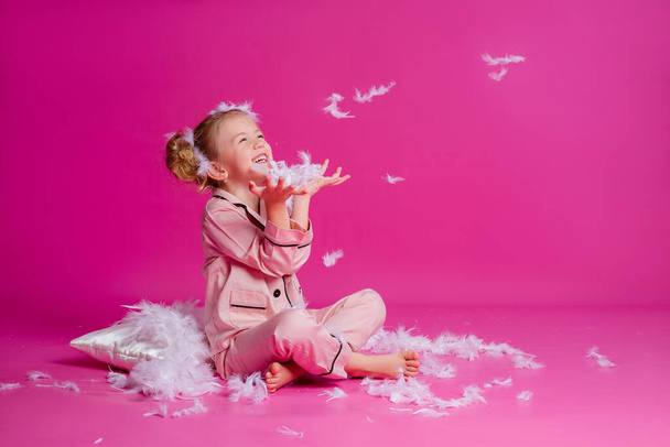 Photo studio d'une belle petite fille posant en pyjama rose clair. Joyeux bébé fille debout sur un fond rose avec un oreiller, des plumes et une horloge. Concept des émotions des enfants - Photo, image