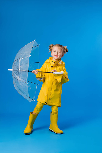 Cheerful girl in a raincoat and rubber boots standing on a blue background with a transparent umbrella. Copy space picture with placement for your text of advertisement - 写真・画像