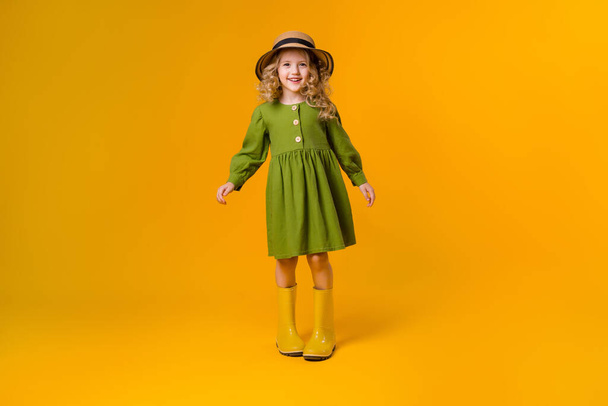 Photo of happy girl in a green linen dress, yellow rubber boots and straw hat on a yellow background - Foto, Imagem