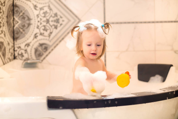Petite fille mignonne se baigner dans le bain avec de la mousse à la maison, joue avec le canard en caoutchouc  - Photo, image