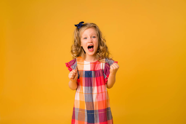 Little curly girl in a plaid check dress on orange background. Children's emotions concept - Fotoğraf, Görsel