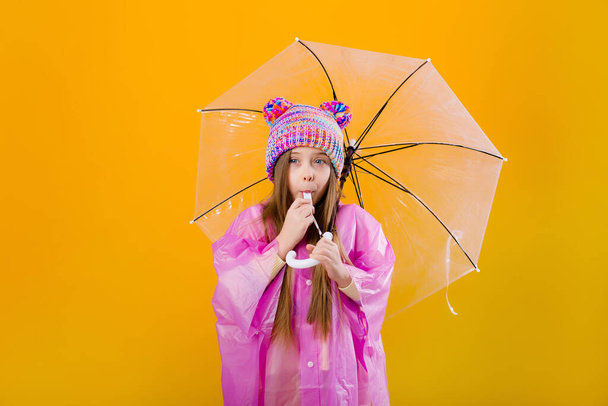 Photo of a little girl in a pink raincoat and  knitted hat holding an umbrella isolated on yellow background with a space for text - Photo, Image