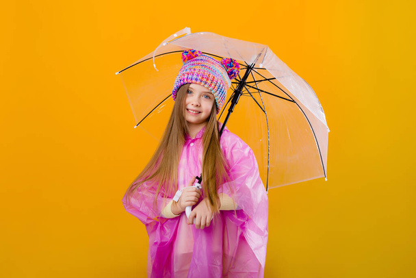 Photo of a little girl in a pink raincoat and  knitted hat holding an umbrella isolated on yellow background with a space for text - Fotografie, Obrázek