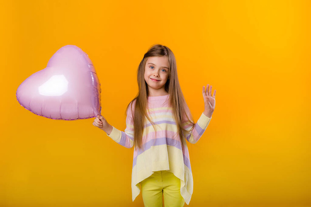 Retrato de una niña feliz sonriendo y sosteniendo un globo rosa en forma de corazón. Aislado sobre fondo amarillo con espacio para texto - Foto, Imagen