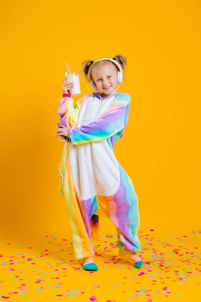 A happy little girl in a unicorn  kigurumi listens to music in headphones from a smartphone. Isolated on yellow background with  space for text - Foto, immagini