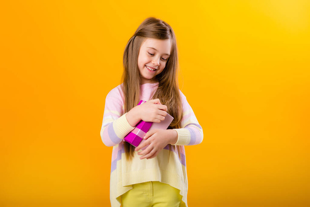 Picture of a happy little girl  holding a pink gift box, isolated on yellow background with a space for text - Foto, Bild