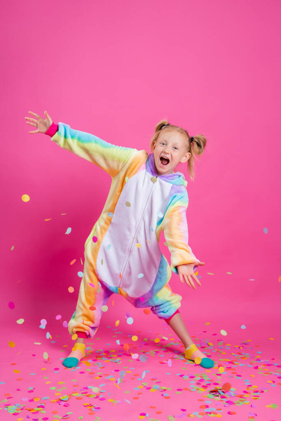 Boldog kislány egyszarvú uniformisban rózsaszín háttérrel szórakozik sokszínű konfettivel. Rózsaszín alapon elszigetelve, szöveges szöveggel - Fotó, kép