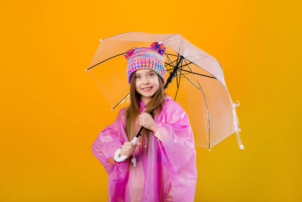 Photo of a little girl in a pink raincoat and  knitted hat holding an umbrella isolated on yellow background with a space for text - Foto, imagen