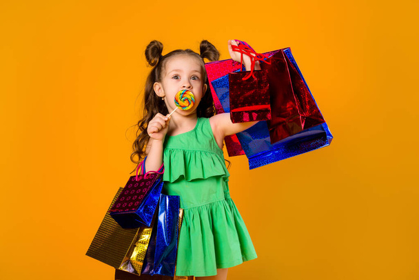 Cute charming girl with lollipop and colorful shopping bags on a yellow background. Copy space picture with placement for your text or advertisement - Photo, image