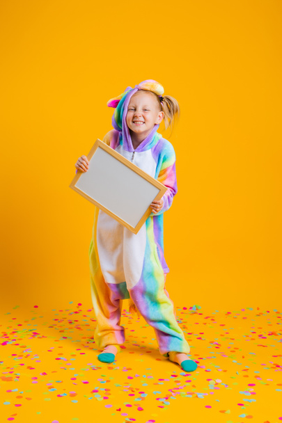 Una niña feliz en un unitard unicornio kigurumi sostiene un tablero vacío para el texto sobre un fondo amarillo. Concepto de vacaciones o información sobre descuentos - Foto, imagen