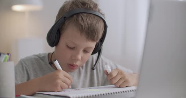 engraçado menino da escola está ouvindo professor em fones de ouvido durante a sessão de aprendizagem remota e desenho de imagem em Copybook - Filmagem, Vídeo