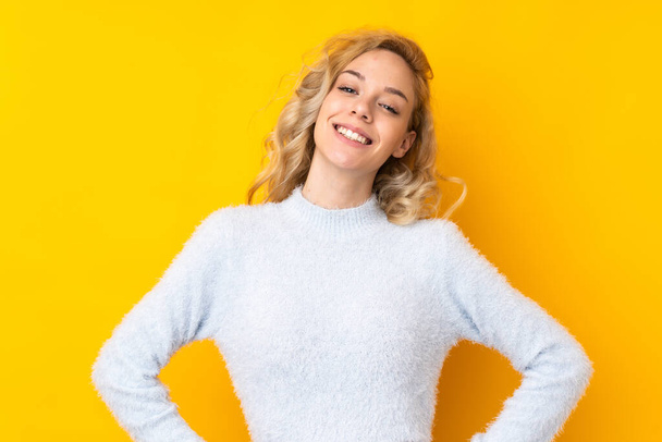 Young blonde woman isolated on yellow background posing with arms at hip and smiling - Фото, изображение