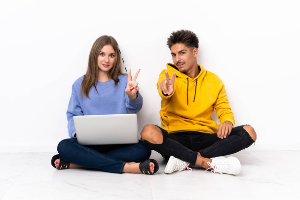 Young couple with a laptop sitting on the floor isolated on white background smiling and showing victory sign - Fotoğraf, Görsel