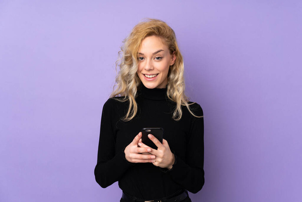 Young blonde woman isolated on purple background sending a message with the mobile - Fotografie, Obrázek