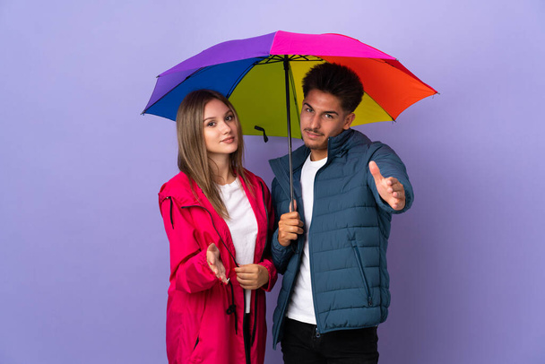 Couple holding an umbrella isolated on blue background shaking hands for closing a good deal - 写真・画像
