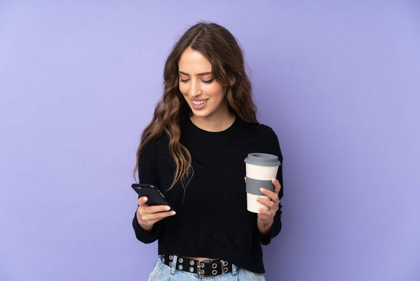 Jonge vrouw over geïsoleerde paarse achtergrond houden koffie mee te nemen en een mobiele - Foto, afbeelding