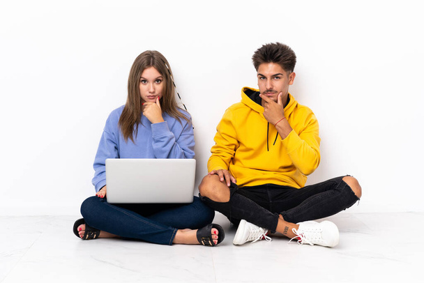 Young couple with a laptop sitting on the floor isolated on white background thinking - Фото, зображення