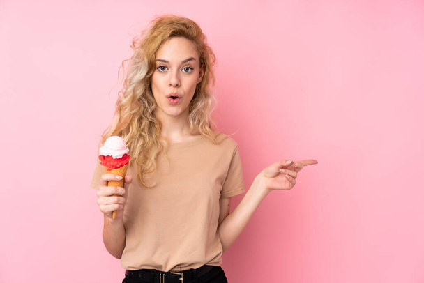 Giovane donna bionda in possesso di un gelato alla cornetta isolato su sfondo rosa sorpreso e indicando lato - Foto, immagini