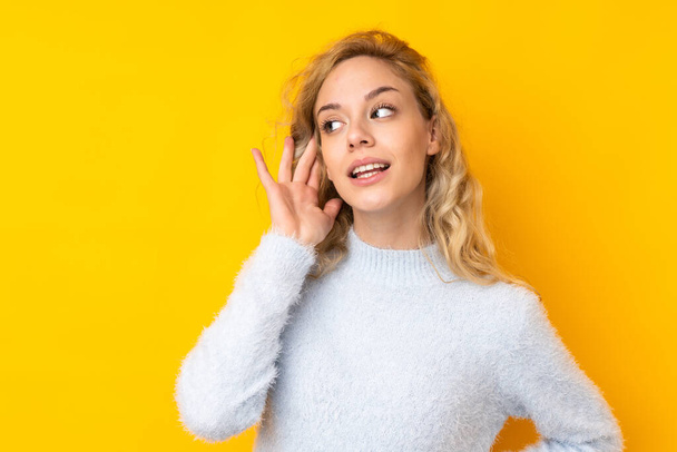 Young blonde woman isolated on yellow background listening to something by putting hand on the ear - Φωτογραφία, εικόνα