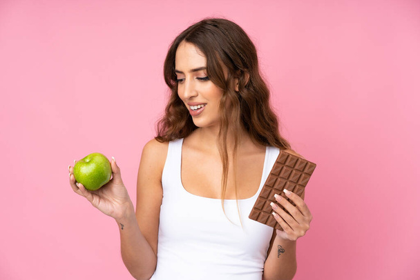 Молодая женщина на изолированном розовом фоне берет шоколадную таблетку в одной руке и яблоко в другой - Фото, изображение