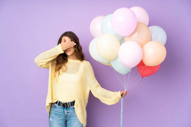 Junge Frau fängt viele Luftballons über isoliert auf lila Hintergrund bedeckt Augen mit den Händen - Foto, Bild