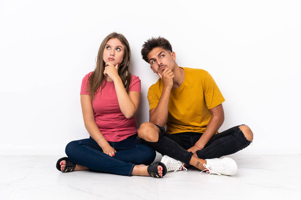 Young couple sitting on the floor isolated on white background having doubts - Photo, Image