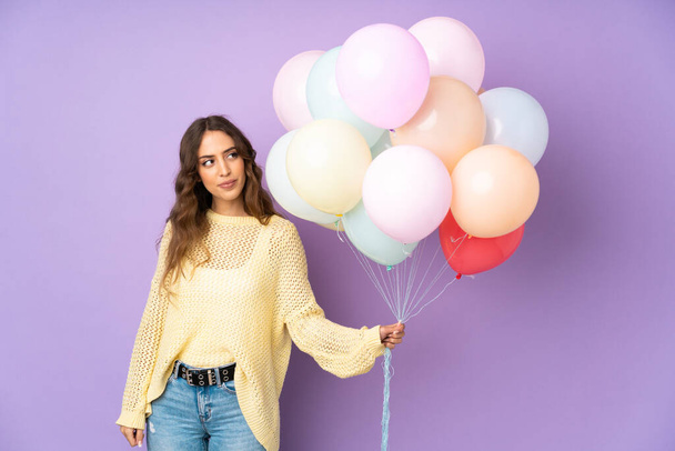 Młoda kobieta łapie wiele balonów na odizolowanych na fioletowym tle stojąc i patrząc w bok - Zdjęcie, obraz