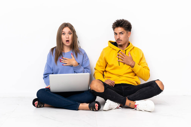 Молода пара з ноутбуком, що сидить на підлозі ізольовано на білому тлі, здивована і шокована, дивлячись праворуч
 - Фото, зображення