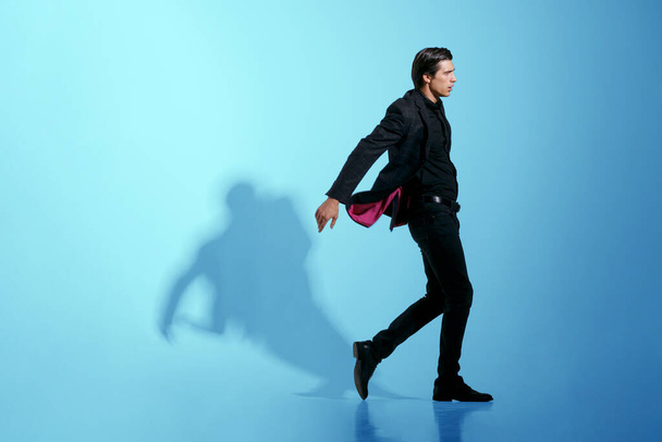 Εικόνα κίνησης ενός ελκυστικού ισχυρού νεαρού άνδρα με πλήρως κομψό μαύρο κοστούμι, απομονωμένο σε μπλε φόντο. - Φωτογραφία, εικόνα