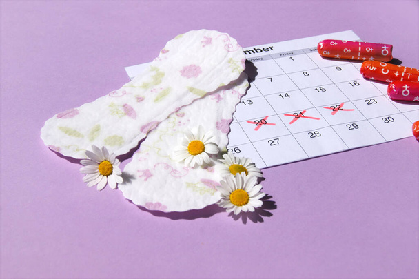 Menstrual pads and tampons on menstruation period calendar with chamomiles on pink background. The concept of female health, personal hygiene during critical days. - Photo, Image