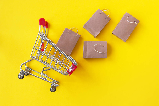 Mini carrito de compras con pequeñas bolsas de artesanía en el fondo amarillo. Vista superior. El concepto de compras en tiendas y en línea. - Foto, imagen