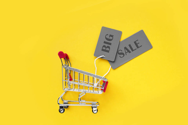 Mini carrito de compras con etiquetas grises con una inscripción GRANDE VENTA sobre un fondo amarillo. El concepto de compras en tiendas y en línea. - Foto, imagen