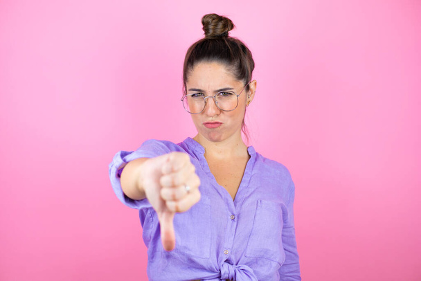 Jeune belle femme portant des lunettes sur fond rose isolé avec le visage en colère, signe négatif montrant aversion avec le pouce vers le bas - Photo, image