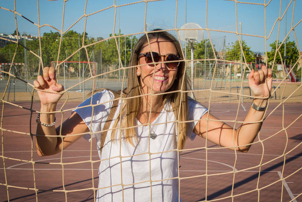 Σύγχρονη γυναίκα γελάει. Νεαρή γυναίκα που γελάει ενάντια στο φράχτη του μπάσκετ. Αστεία γυναίκα ντυμένη στα λευκά και φορώντας γυαλιά ηλίου. Τρόπος ζωής - Φωτογραφία, εικόνα