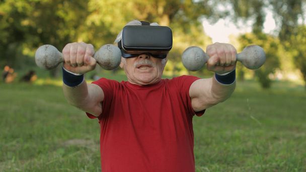 Anciano abuelo en casco VR haciendo ejercicios de fitness con pesas en el parque - Foto, imagen