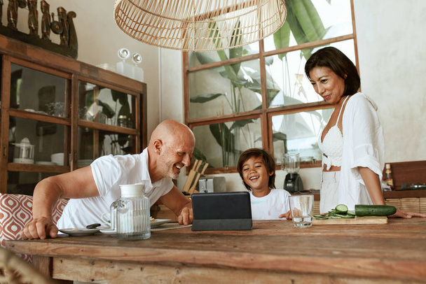 Family Cooking. Happy Parents With Kid Using Tablet At Kitchen Enjoying Weekend. Healthy Food. Positive Mother, Father And Son On Summer Vacation At Tropical Resort. Vegetarian Diet As Lifestyle. - Photo, Image