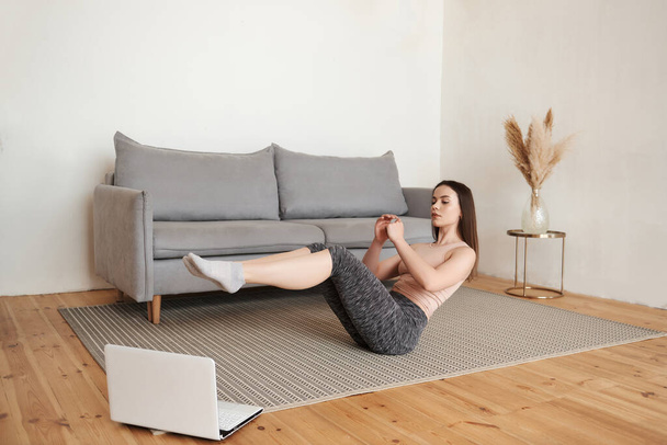 Girl watching videos on laptop and repeating online instructions, doing abdominal exercises for upper and lower abs on the floor, doing v-ups abs workout in the living room. Copy space, full length. - Photo, Image