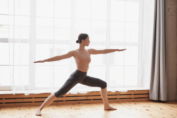 Junge Frau übt Yoga, Stretching-Übungen, steht in Kriegerpose zu Hause oder in einem Yogastudio mit großem Fenster, Kopierraum, Seitenansicht, waagerecht, trägt Sportbekleidung, drinnen in voller Länge - Foto, Bild