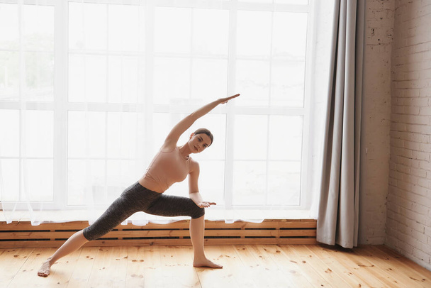 Young woman practice yoga, stretching in Bending Side exercise, working out indoor in yoga studio with a big window. Healthy lifestyle, yoga concept, wearing sportswear, pants and top, full length - 写真・画像