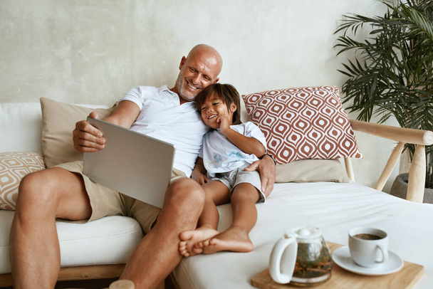 Father And Son With Tablet Portrait. Mixed Race Family Of Dad And Little Boy Using Portable Digital Device Sitting On Sofa Together. Technology For Education And Fun At Tropical Resort. - 写真・画像