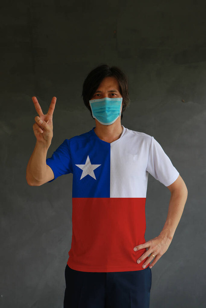 Мужчина в маске одет в рубашку цвета флага Чили и поднимает два пальца на темном фоне стены. Концепция защиты и борьбы с COVID. - Фото, изображение
