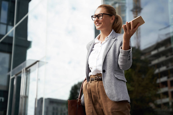 Счастливая успешная деловая женщина в очках и классической одежде держит мобильный телефон и улыбается стоя напротив офисного здания на открытом воздухе - Фото, изображение