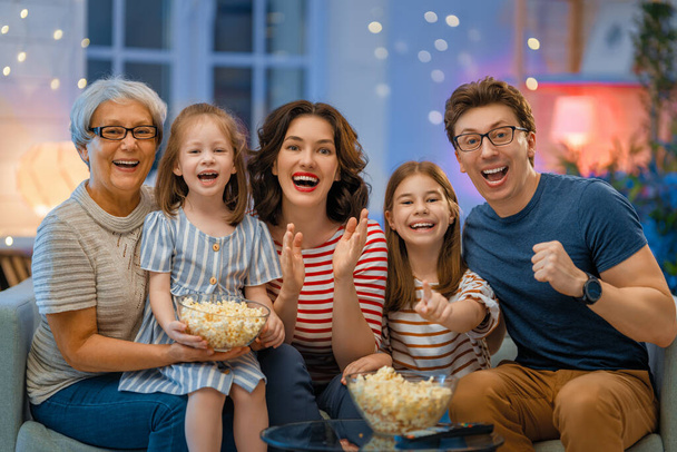 Happy family watching projector, TV, movies with popcorn in the evening at home. Granny, mother, father and daughters spending time together. - 写真・画像