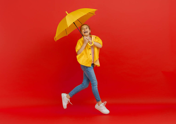 Happy emotional child laughing and jumping. Kid with yellow umbrella on colored red background. - Фото, изображение