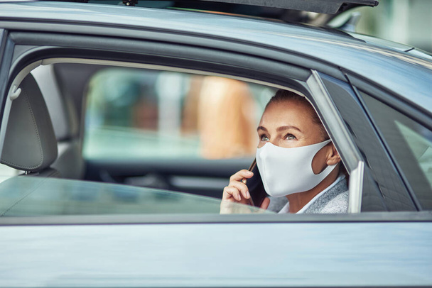 Üzlet, mint új normális koncepció, oldalnézet egy fehér üzletasszony orvosi védőmaszkot visel beszél okostelefonon, miközben ül a hátsó ülésen a kocsiban - Fotó, kép