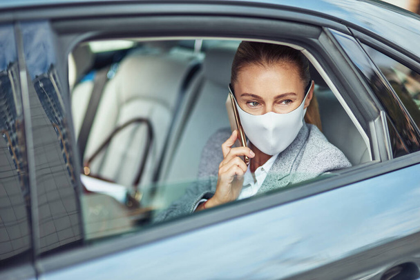 Ковид 19 или вспышка коронавируса, белая деловая женщина в медицинской защитной маске, разговаривающая по смартфону, сидя на заднем сидении в машине, глядя в окно - Фото, изображение